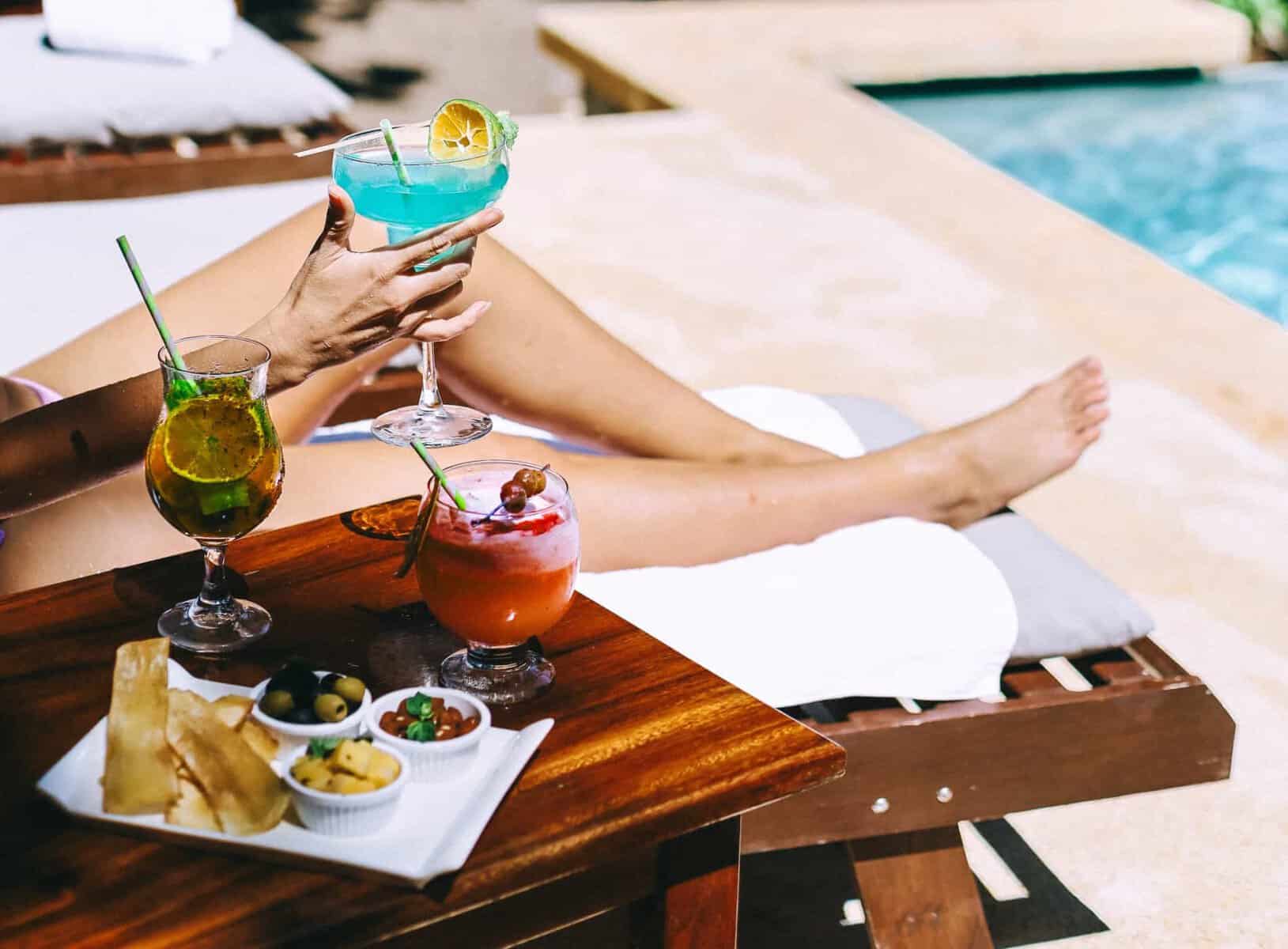 A woman is sitting next to a pool with drinks and food.