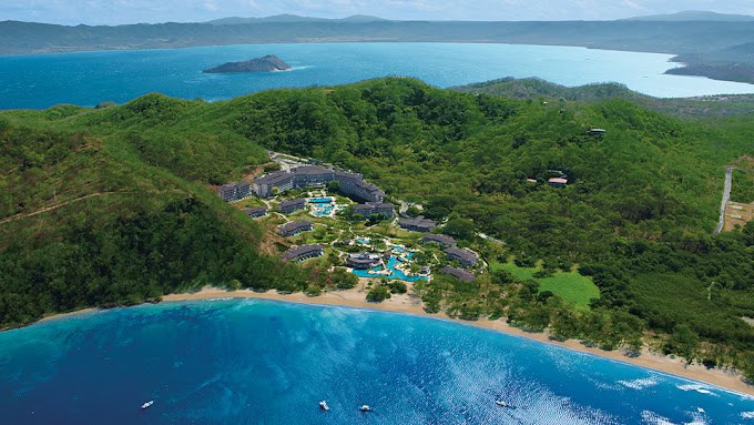 Aerial view of a tropical resort with multiple swimming pools nestled between lush green hills and a clear blue ocean.