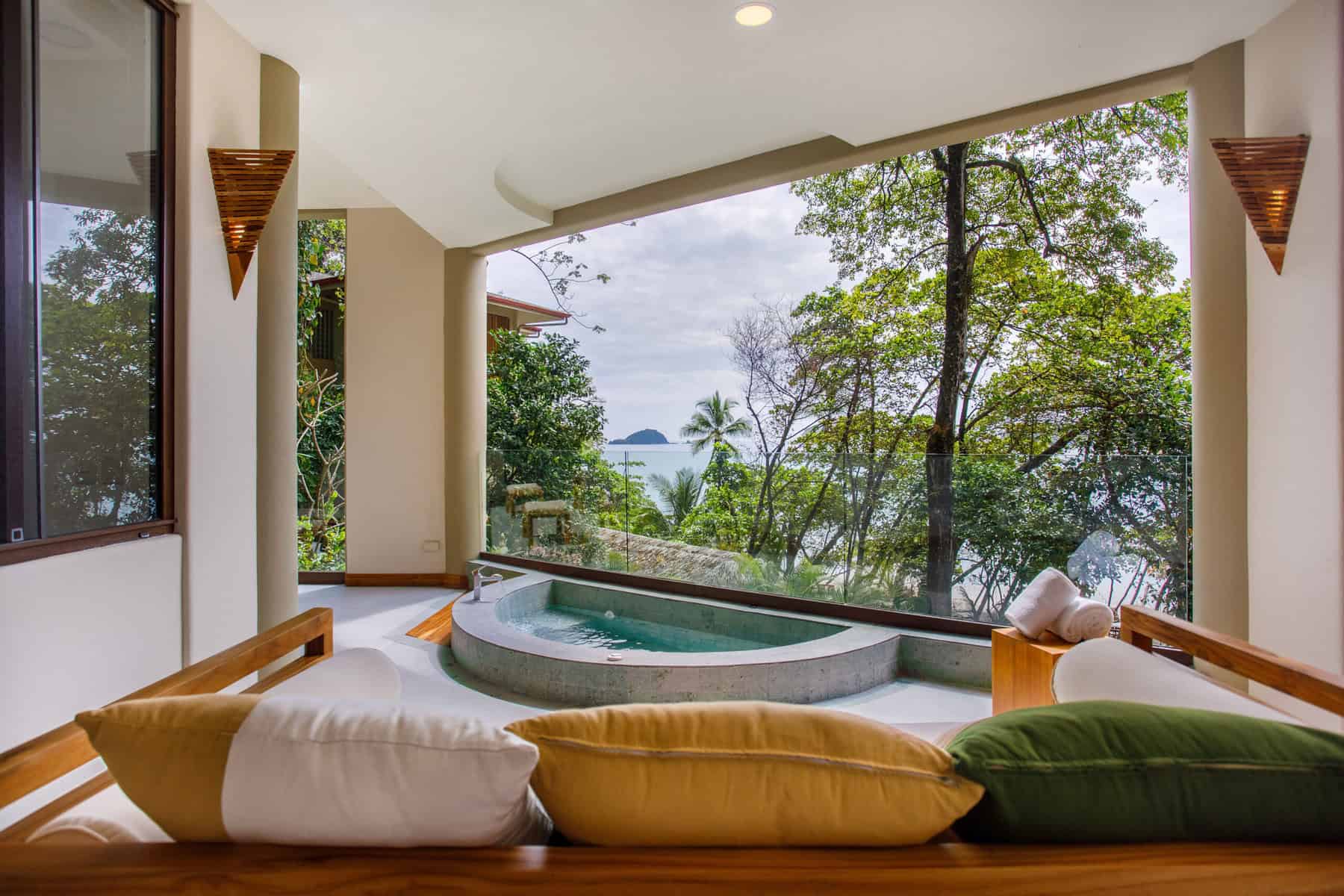 Luxurious room with a view of the sea through the trees, featuring a cozy sitting area and an indoor hot tub.