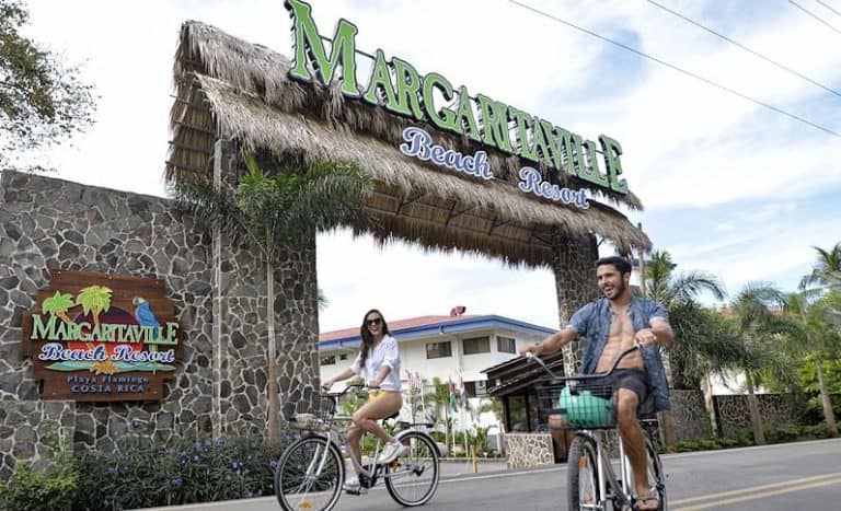 A man and a woman riding bicycles past the entrance of margaritaville beach resort, featuring a large sign with a thatched roof.