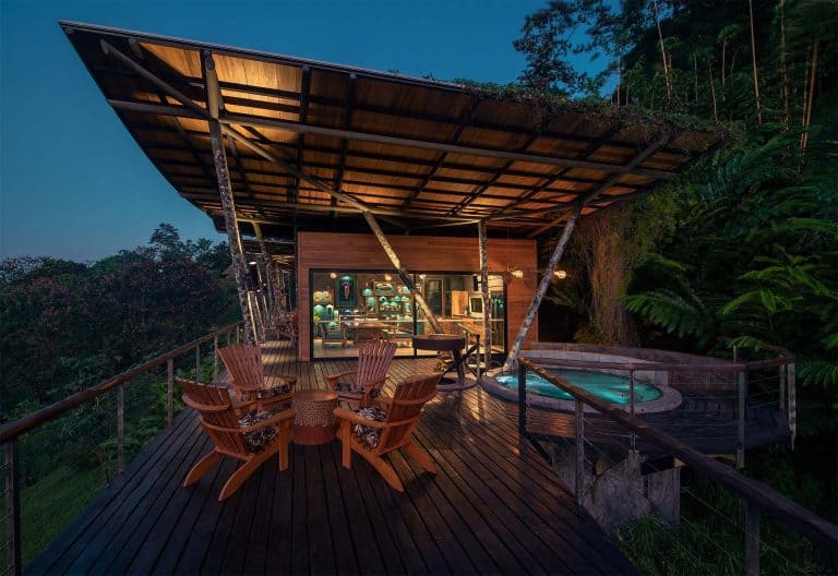 Open-air wooden deck with a seating area and hot tub overlooking a lush forest at dusk.