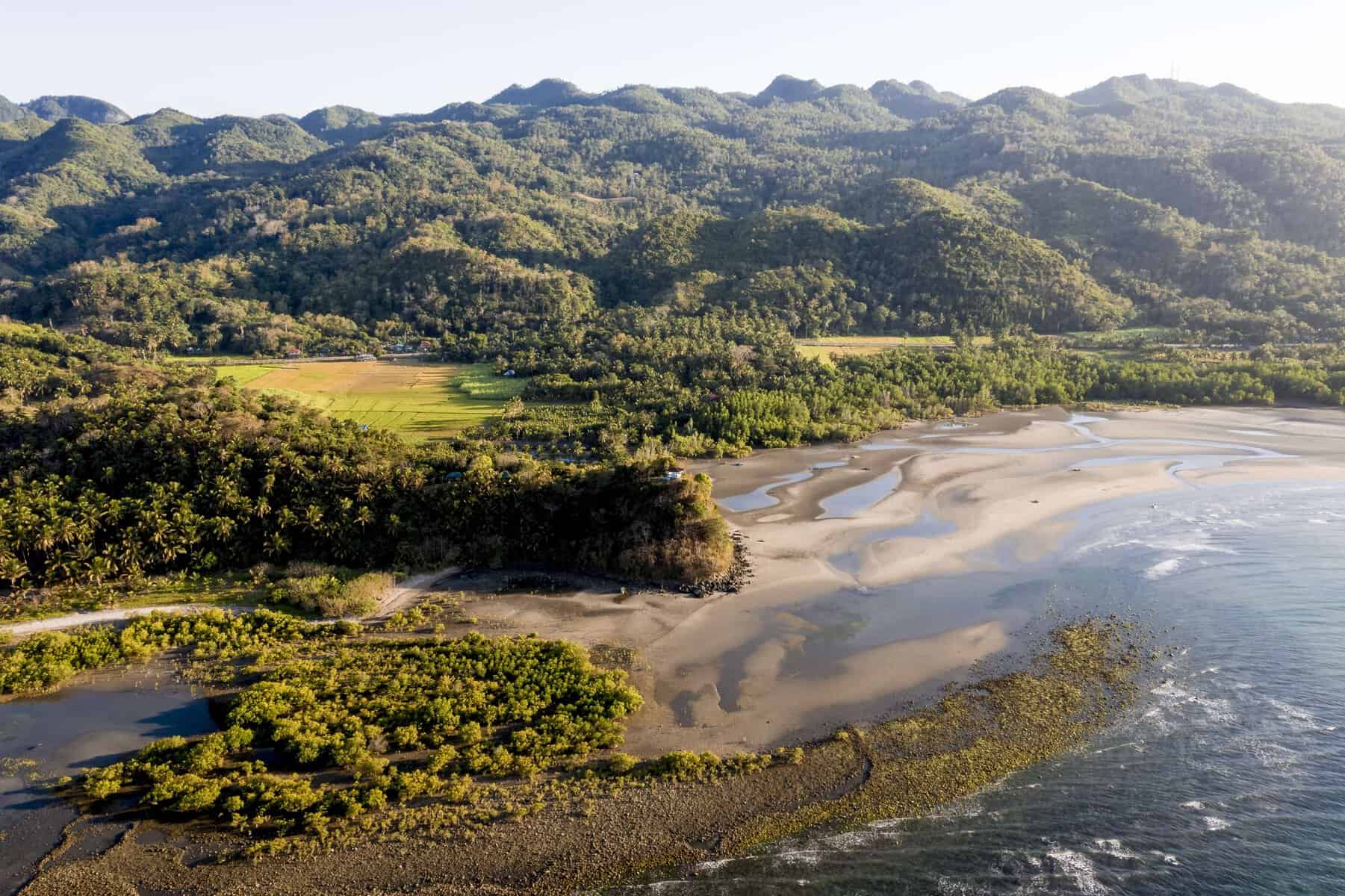 Aerial view of a coastal landscape featuring a stretch of beach, shallow waters, lush greenery, and mountainous terrain in the background.