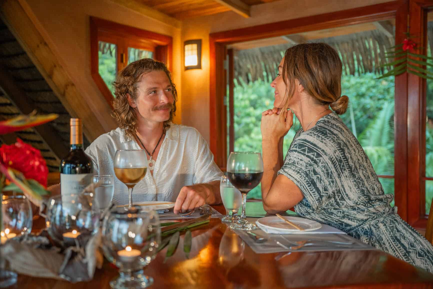 A man and woman sit at a candlelit table with wine and water, talking and smiling in a cozy, tropical restaurant setting.