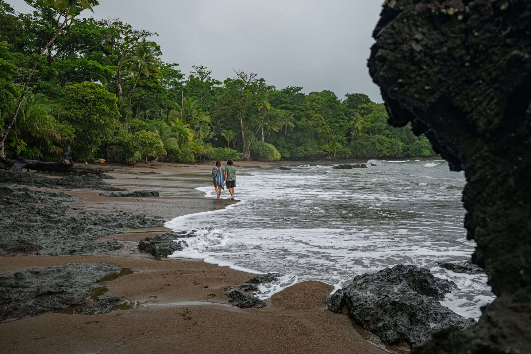 Two people walk along a rocky, tree-lined beach with gentle waves under an overcast sky.