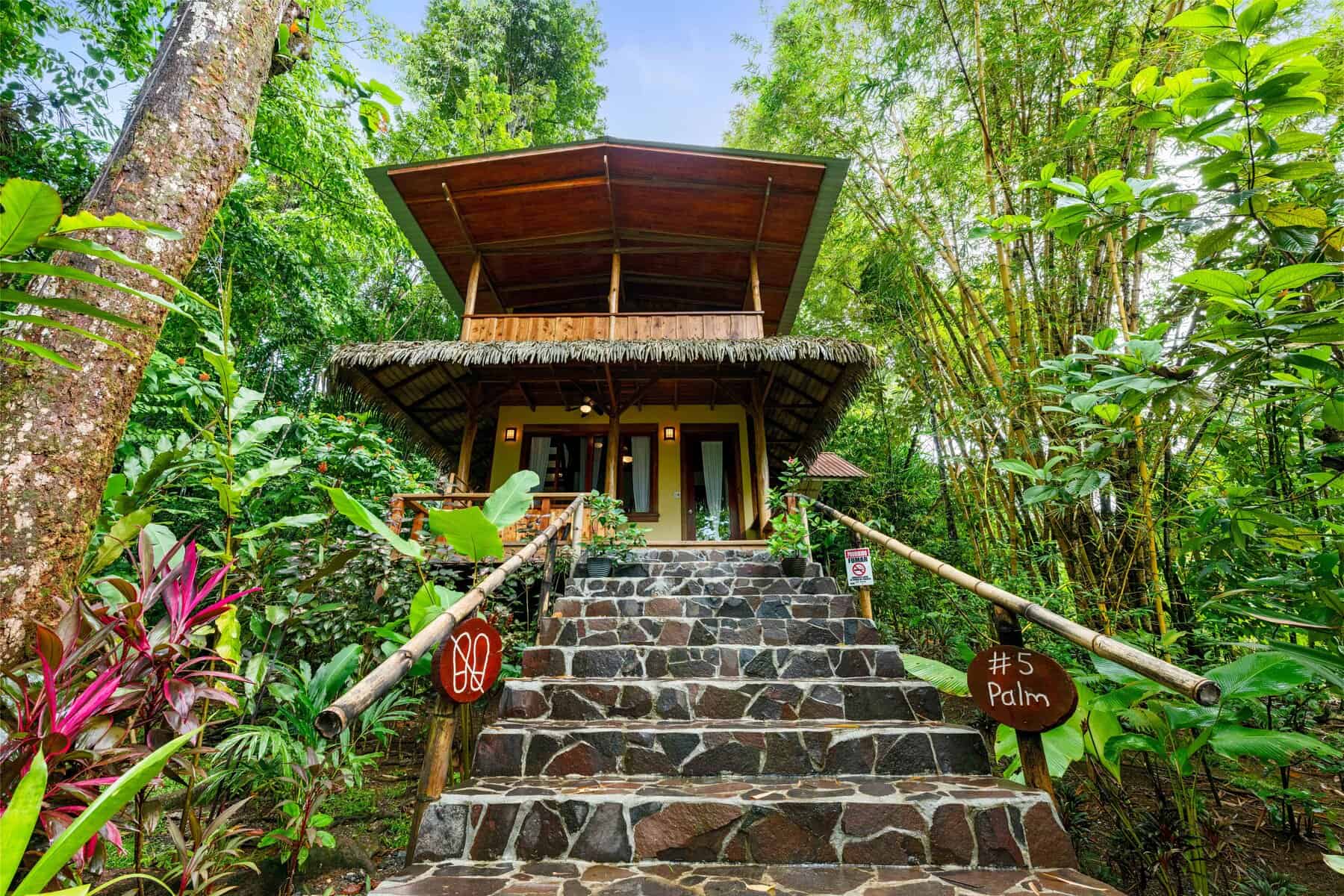 A two-story wooden cabin with a thatched roof sits among lush tropical plants, stone steps leading to the entrance, and a sign reading "#5 Palm" at the base.