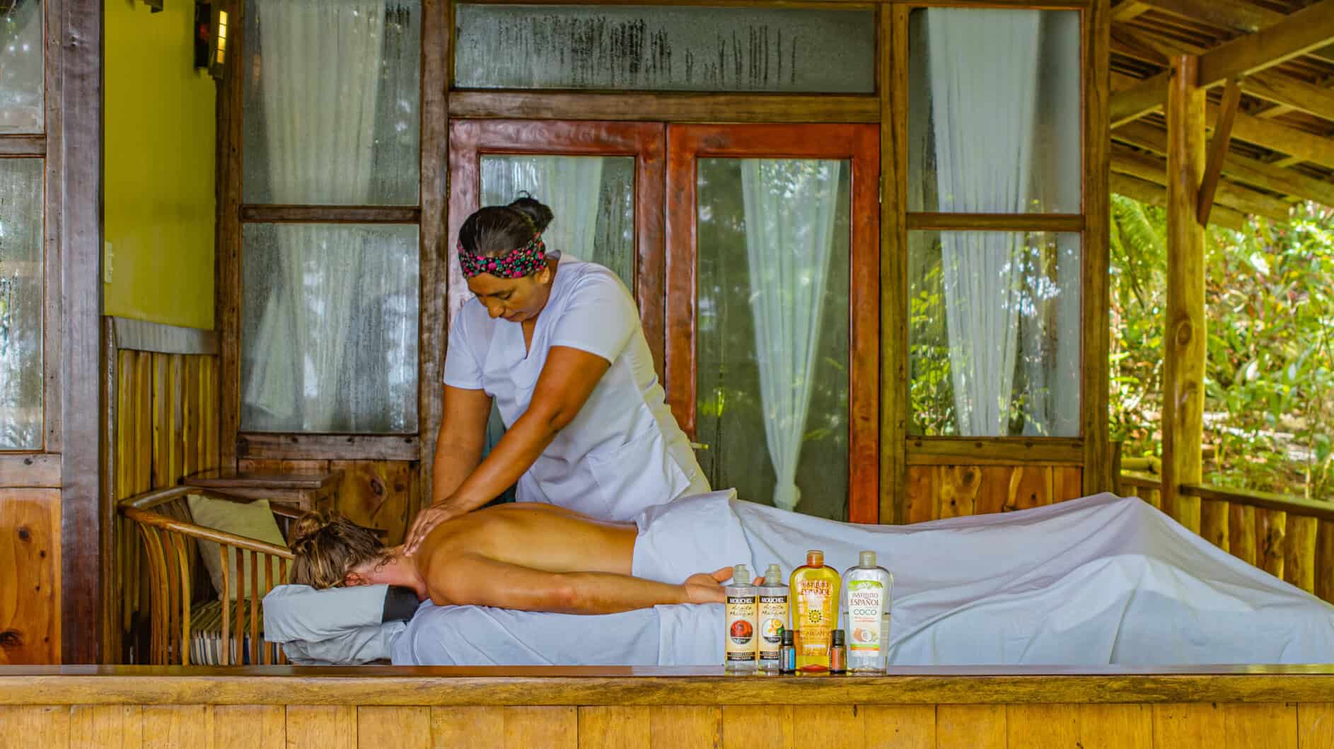 A massage therapist in uniform gives a back massage to a person lying face down on a table in an outdoor wooden spa, with massage oils displayed in the foreground.