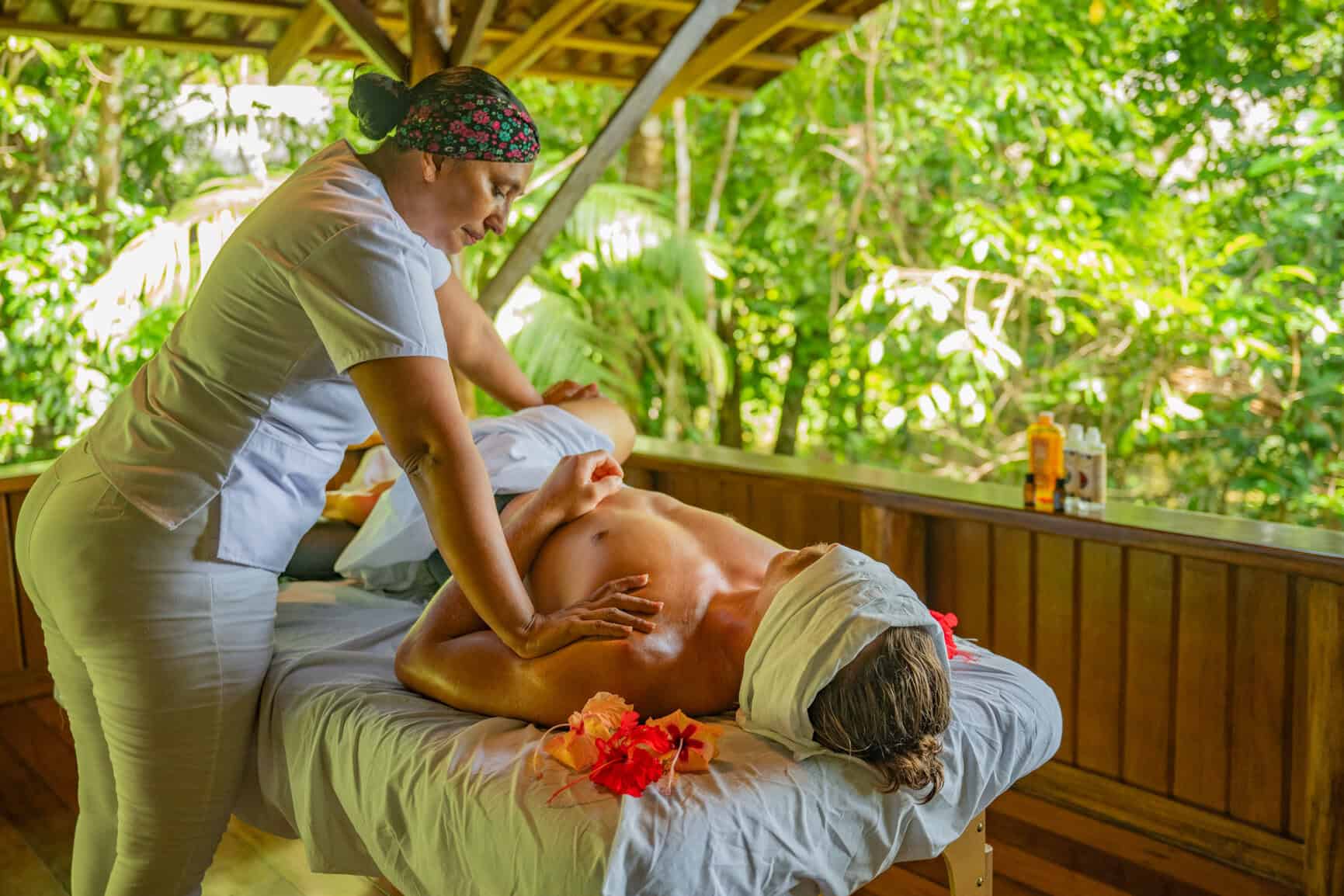 A person receives a massage from a therapist on an outdoor table surrounded by lush greenery; flower petals decorate the table.