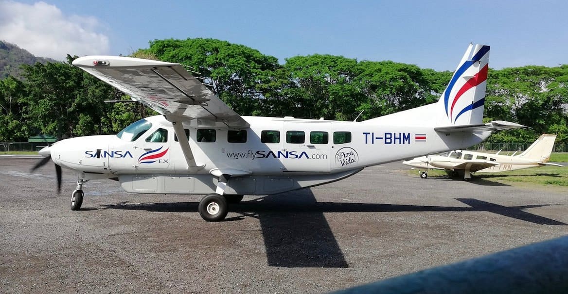 A small white SANSA aircraft with the registration TI-BHM is parked on a tarmac next to another light plane, with trees and a hill in the background.