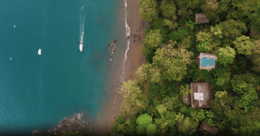 Aerial view of a tropical coastline with turquoise water, boats, sandy beach, rocky shore, and several houses surrounded by dense green forest—an ideal setting for the best Costa Rica hotel experience.