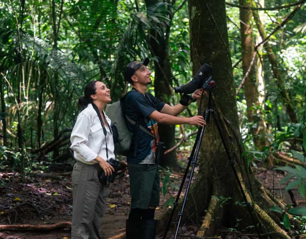 Two people in outdoor clothing stand in a dense forest near the Best Costa Rica Hotel, one adjusting a camera on a tripod while the other looks on, both appearing focused on the lush surroundings.