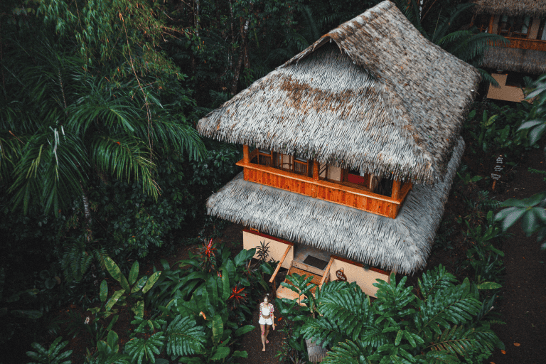 A woman standing in front of a hut surrounded by trees.