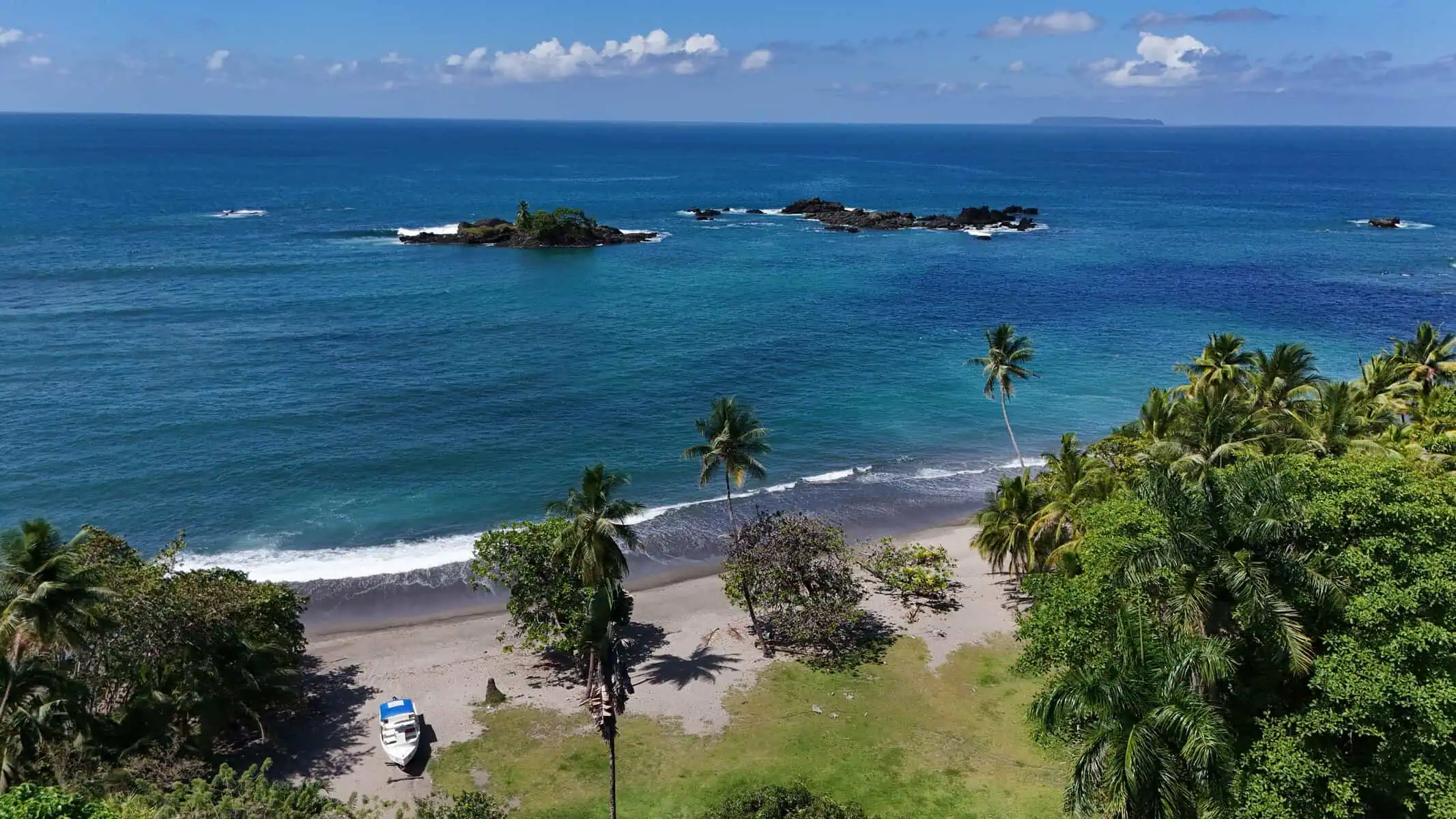 Aerial view of a tropical beach with palm trees, a parked white vehicle, dark sand, and small rocky islands in the blue ocean under a partly cloudy sky.