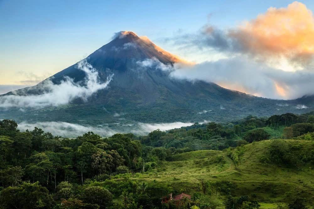 A tall, conical volcano with a cloud-covered peak rises above dense green forest and grassy hills at sunrise or sunset.