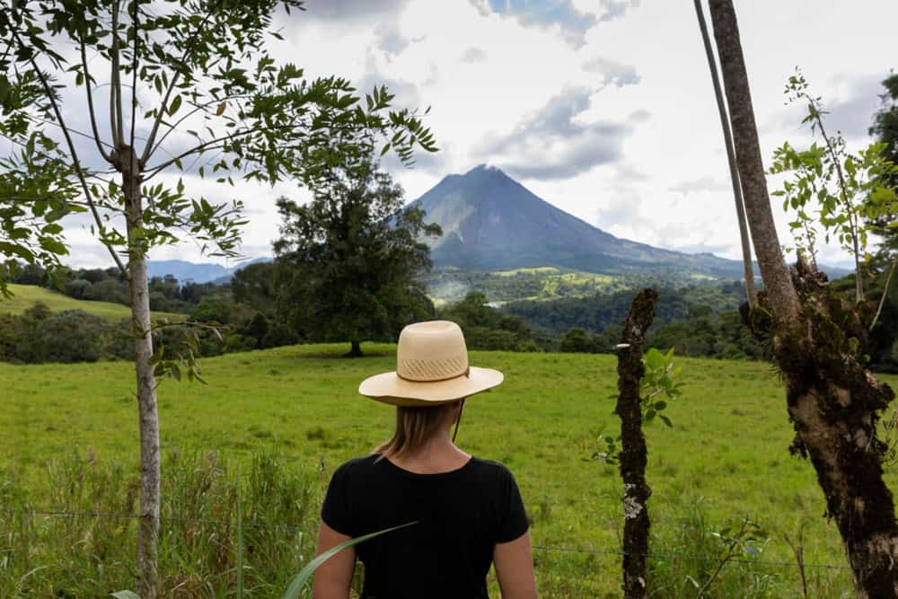 A person wearing a straw hat stands in a grassy field, facing a distant volcanic mountain with lush trees and clouds in the background.