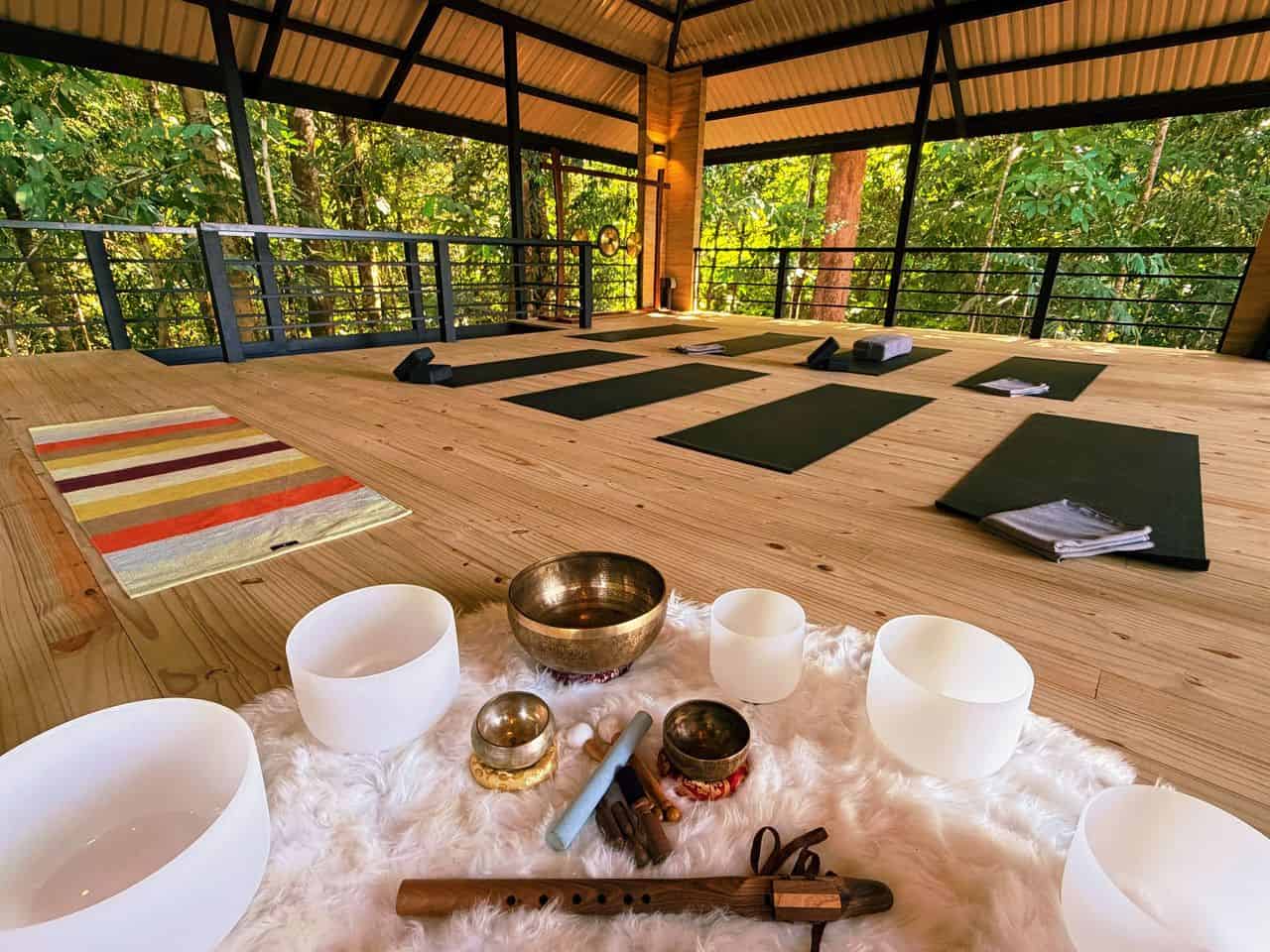 Open-air yoga studio with mats, blocks, and blankets on a wooden floor; singing bowls and instruments arranged on a white rug in the foreground, surrounded by trees.