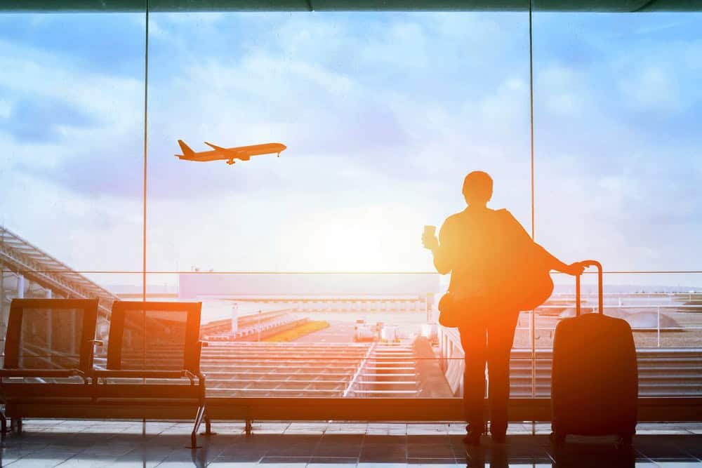 A person stands by an airport window with luggage and a drink, watching an airplane take off against a bright sky.