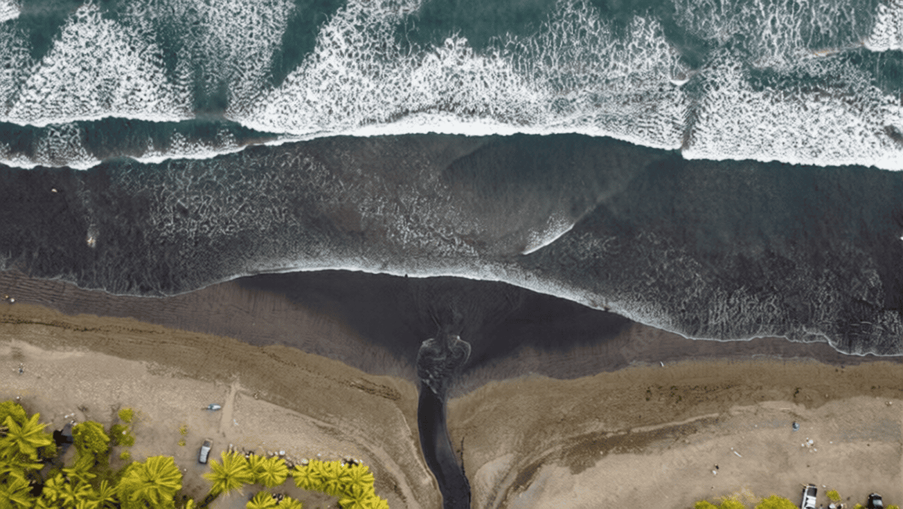 Aerial view of waves breaking on a sandy beach, with a narrow river or stream flowing from the shore into the ocean, bordered by palm trees.