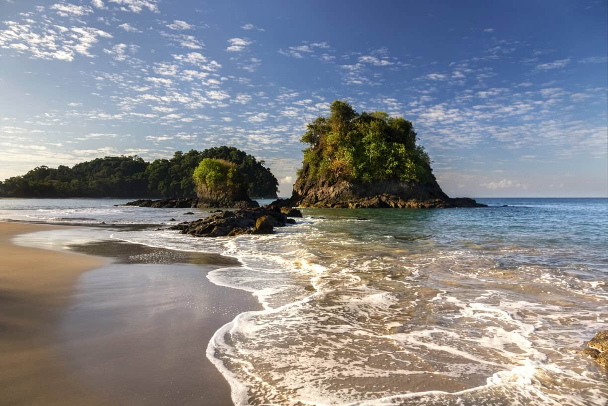 Small rocky islets covered with green vegetation sit close to a sandy beach as gentle waves wash ashore under a partly cloudy sky.