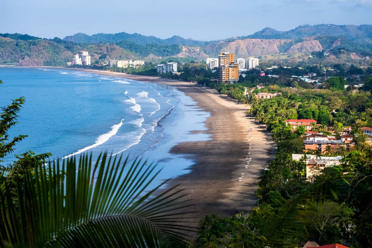 A wide sandy beach curves along the coastline with gentle waves, bordered by buildings, greenery, and distant mountains under a clear sky.