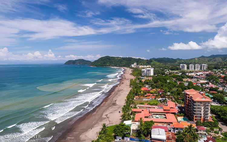 Aerial view of a coastal city with tall buildings, sandy beach, ocean waves, and green hills in the background under a partly cloudy sky.