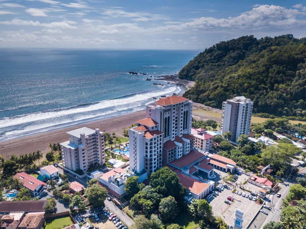 Aerial view of a beachfront hotel complex with several tall buildings, a parking lot, and a forested hill beside the ocean under a partly cloudy sky.