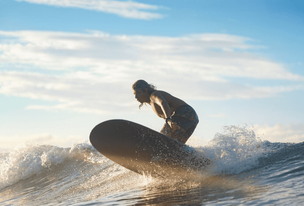 A person rides a wave on a surfboard under a blue sky with scattered clouds, with water splashing around them.
