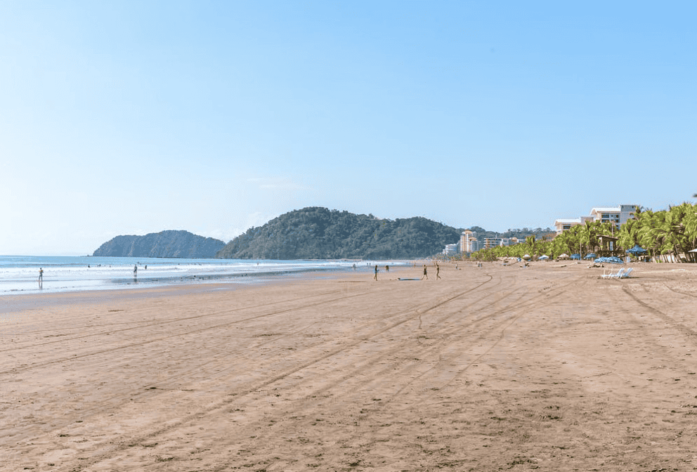 Wide sandy beach with a few people walking, distant hills in the background, and resort buildings with palm trees lining the shore under a clear blue sky.