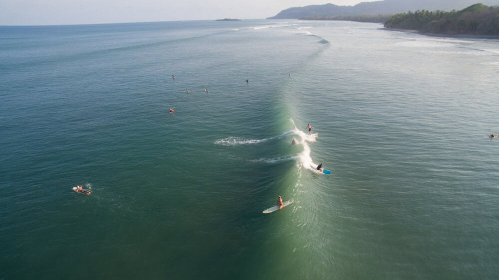 A group of surfers ride and wait for waves in the ocean near a coastline with distant hills and a sandy beach.