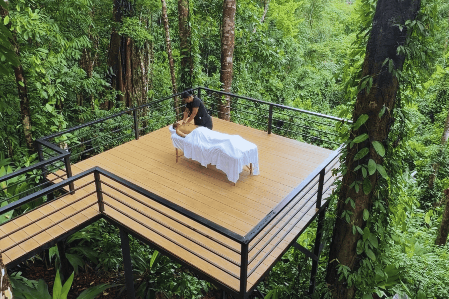 A massage therapist gives a massage to a person lying on a table covered with white sheets on a wooden deck surrounded by lush green forest, creating a tranquil setting reminiscent of yoga on the Osa Peninsula.